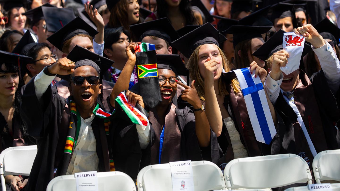Four students at Brown Commencement raising flags and stoles representing their home countries.