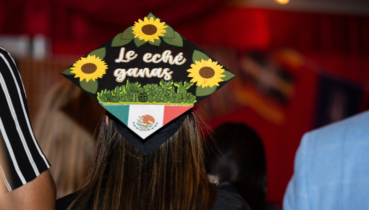 Student wearing a graduation cap decorated with a Mexican flag, sunflowers, and the phrase le eché ganas.