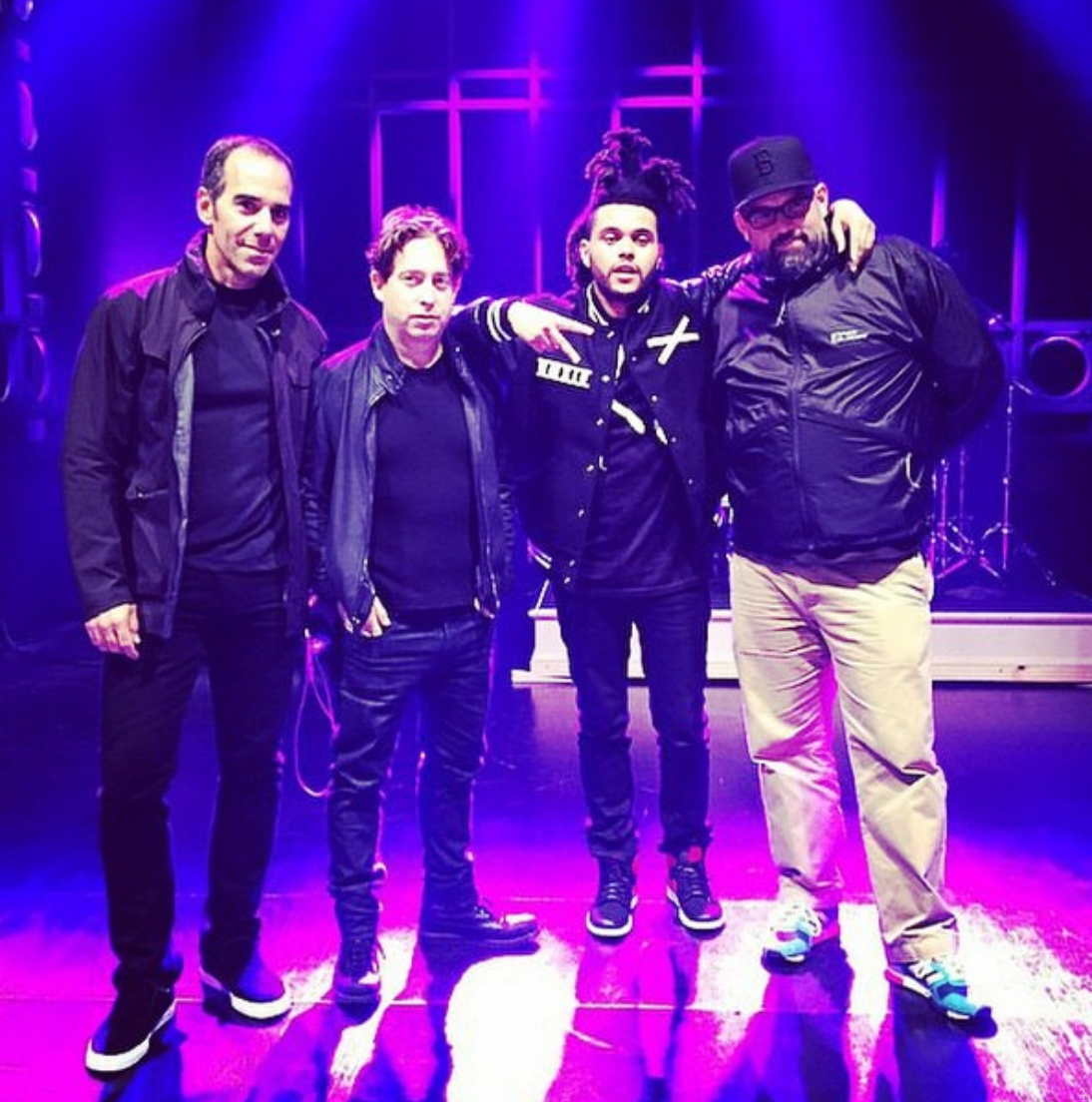 Nate Albert and pop star The Weekend pose in a group photo with two other people on a brightly lit stage.