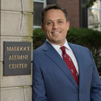 Photo of Carlos Lejnieks posing in front of a plaque reading Maddock Alumni Center