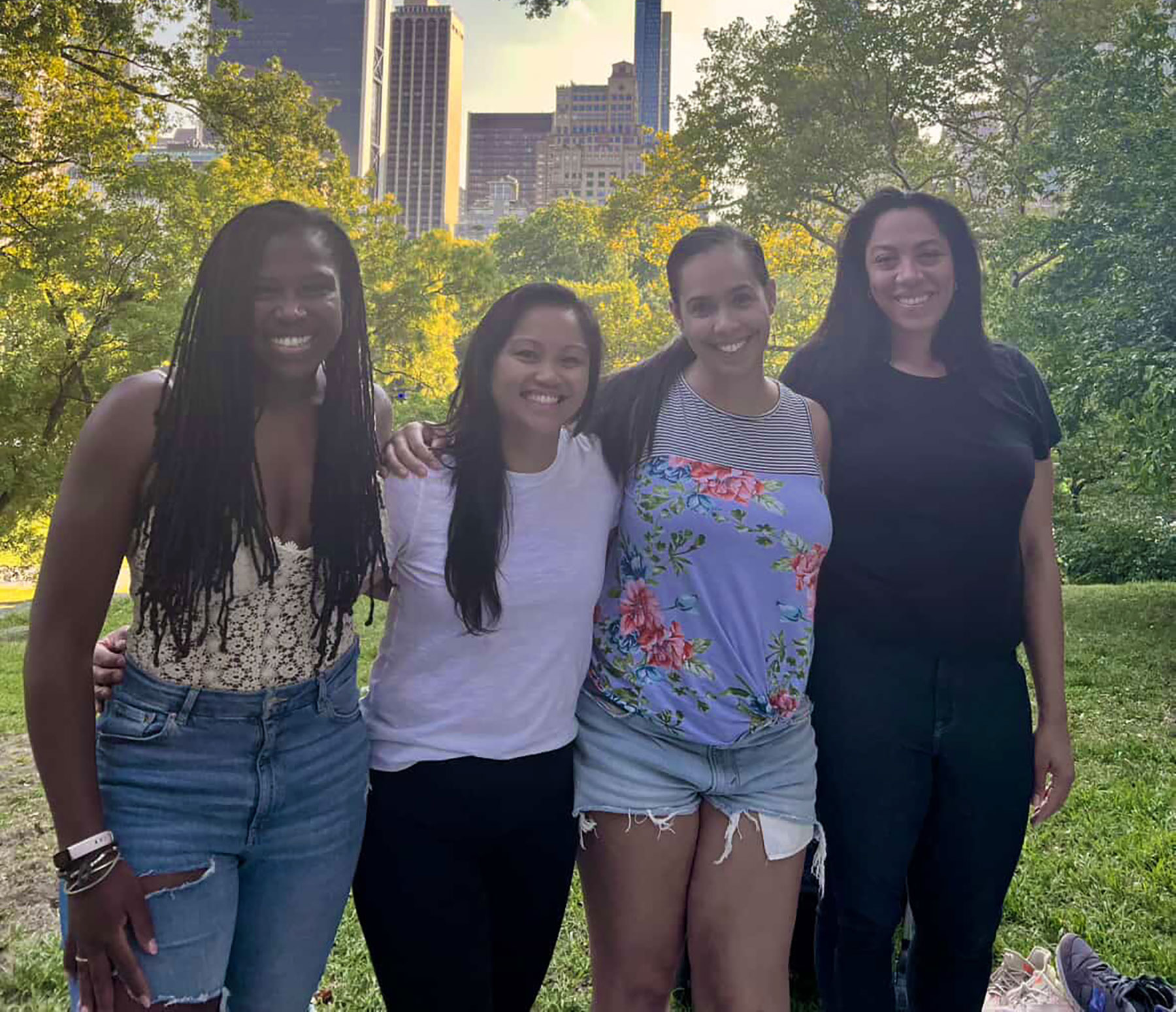 Sheila, Christina, Sthefany, and Aileen are smiling and standing together in a park with a city skyline in the background.