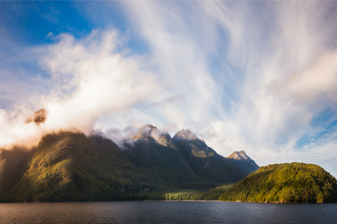 Clouds hang over the green-covered mountain peaks beside placid water in New Zealand.
