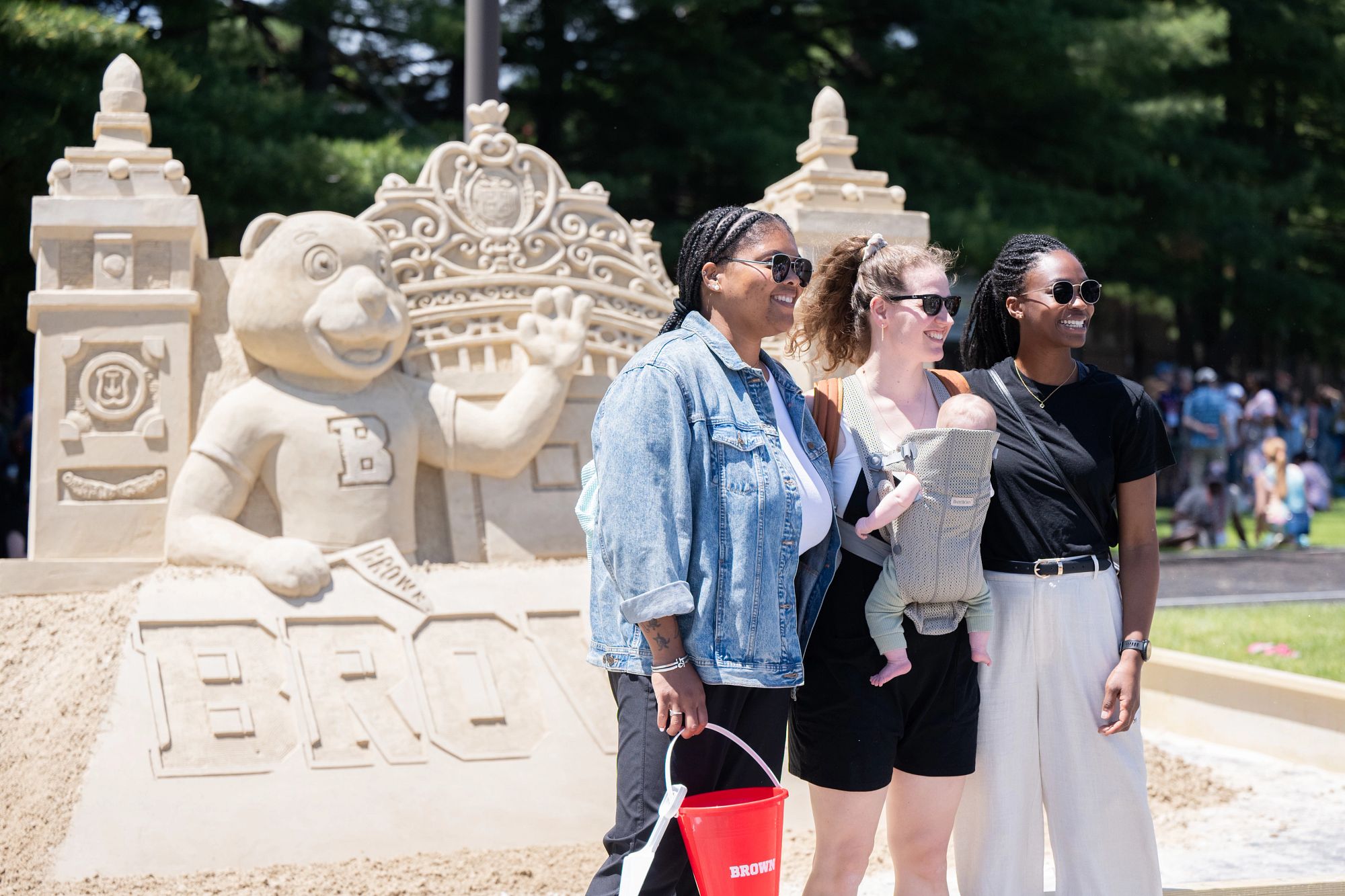 Three women, one with a baby in a carrier on her chest, pose for a photo in front of a sand sculpture of Bruno and the Van Wickle Gates at Bruno's Block Party.
