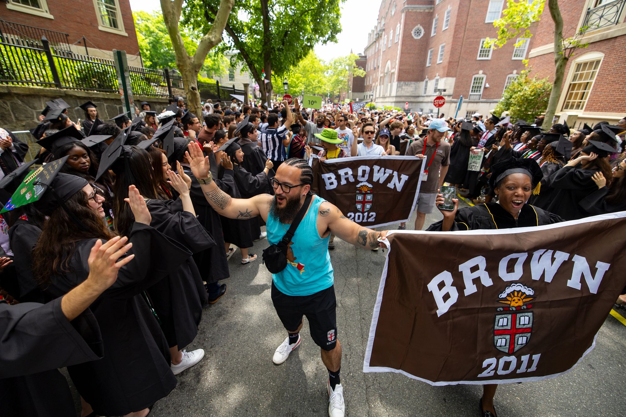 Two alumni hold up a Class of 2011 banner while high-fiving graduates wearing cap and gowns as they walk down College Hill at Procession.