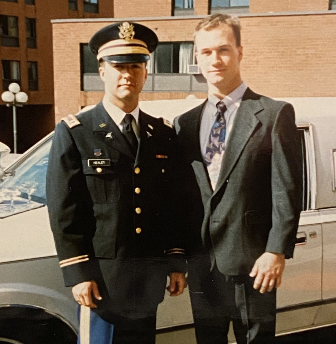 Healey in uniform stands side by side to his brother in front of a car.