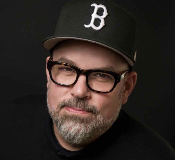 Portrait of Nate Albert wearing glasses and a black baseball cap with the letter “B” embroidered in white. 