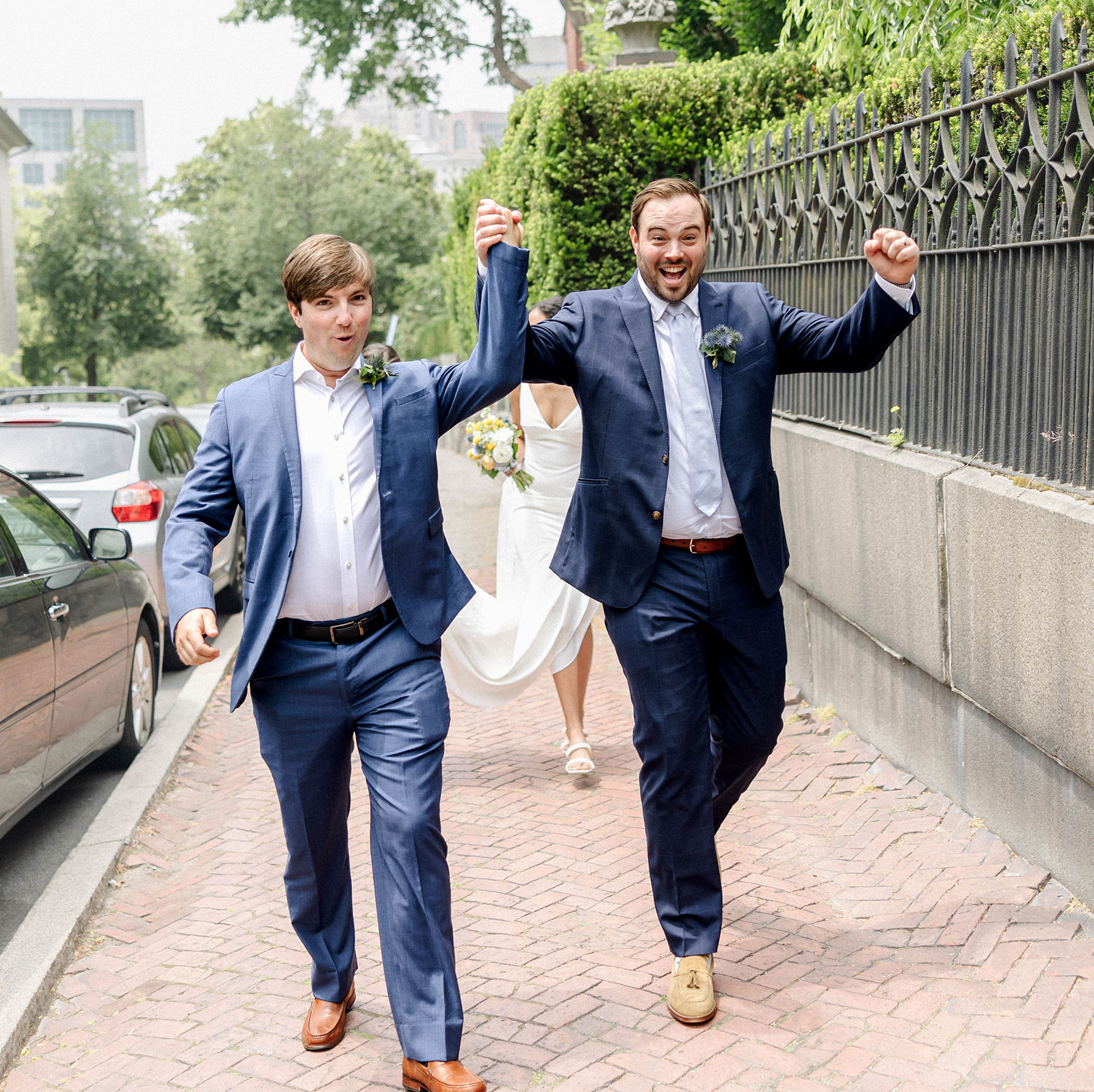 Peter and James in formal attire joyfully walking and holding hands along a city street, with cars and a metal fence beside them.