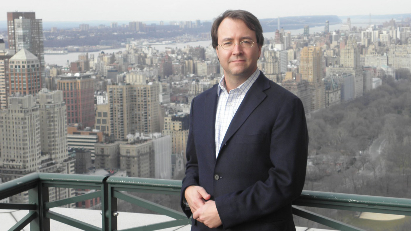 Joe Healey is wearing a suit, posing for a photo with a skyline view of a city behind him.