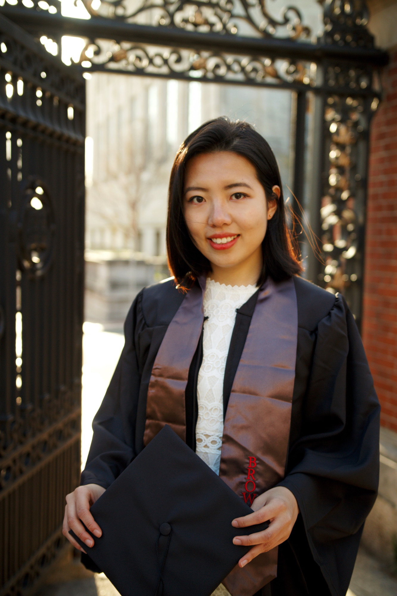 Joy wears a graduation gown and holds a cap while standing near an ornate gate. The background features a blurred cityscape. The text on the graphic reads "BROWN VOLUNTEER" and "Joy Zheng '21".