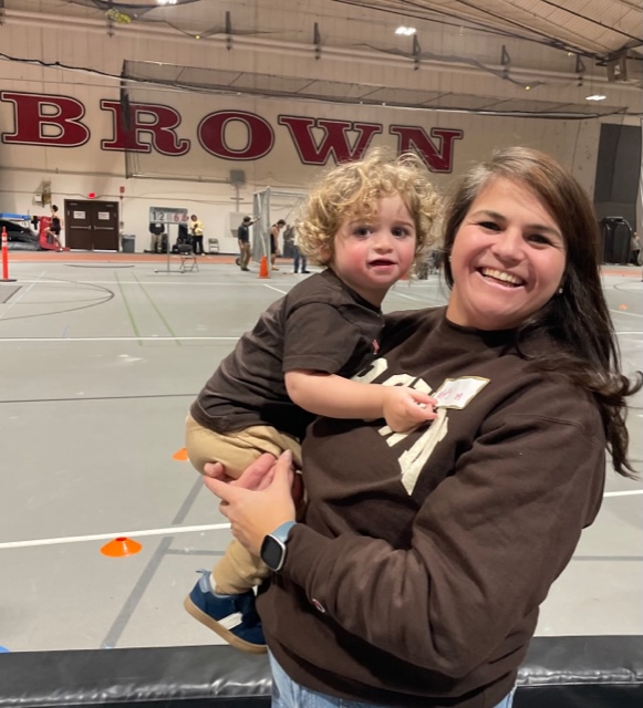 Murphy wearin ga Brown sweatshirt, holding her toddler child in her arms, inside one of Brown's athletic facilities.
