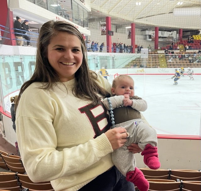 Murphy wearing a Brown sweatshirt and holding a baby in front of Brown's hockey ice rink. 
