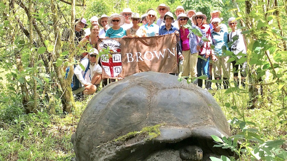 Alumni posing with Brown University banner with tortoise in foreground on Galapagos Islands trip