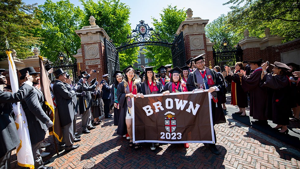 A group of students in graduation attire carrying a Brown banner after walking through the Van Wickle Gates