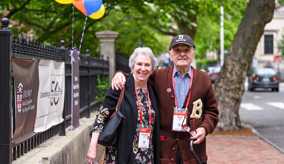 A Brown alum wearing a brown cardigan with a B stitched on the chest and a Brown baseball cap poses outside the gates with a woman.