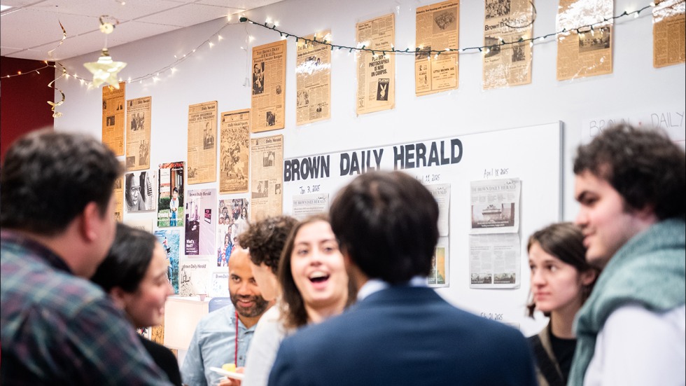 Alums gather inside at the Brown Daily Herald Reunion, with old newspapers displayed on the wall behind them along with string lights.