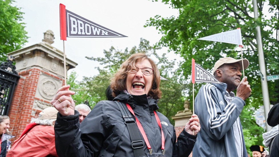 A female alum holding up two Brown pennants smiles joyously as she walks through the Van Wickle Gates during Procession.