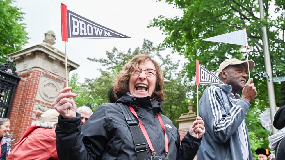 A female alum holding up two Brown pennants smiles joyously as she walks through the Van Wickle Gates during Procession.