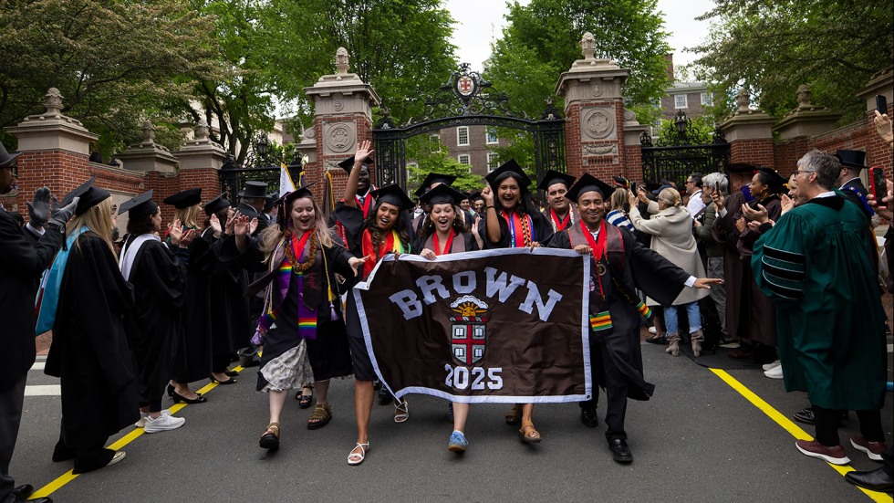 New graduates wearing their caps and gowns hold up their Brown 2025 class banner as they walk through the Van Wickle Gates.