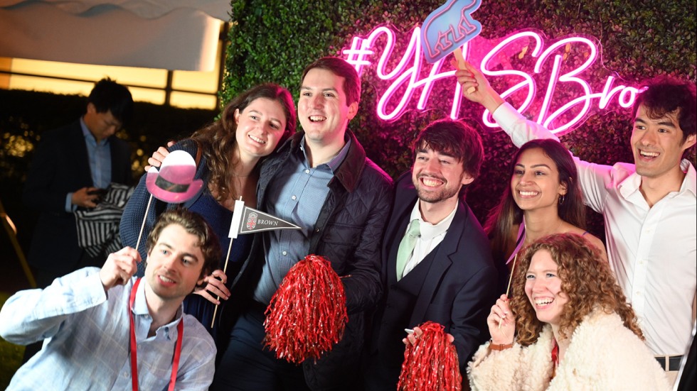A group of young alumni standing in front of a grass backdrop holding props with a pink neon sign reading YASBrown behind them.