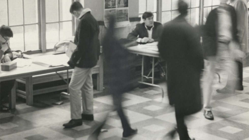 Black and white photo of several individuals in a busy hall in the mailroom, some are in motion and blurred while others are stationary, engaging in activities at tables.