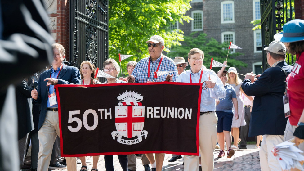 Brown alums celebrating their 50th Reunion hold up their Brown class banner while walking through the Van Wickle Gates at Procession.