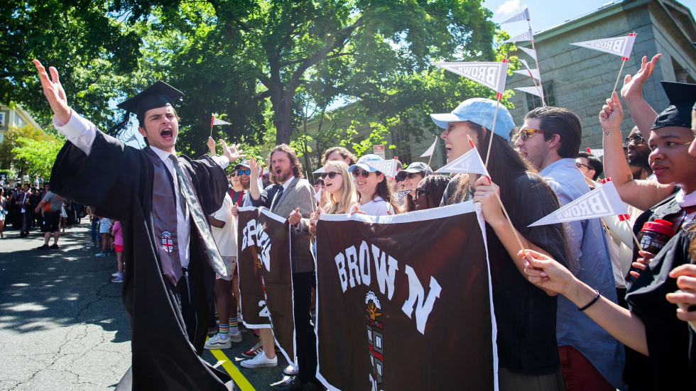 A recent Brown graduate celebrates with a row of alumni lined up on the sidewalk at Procession.
