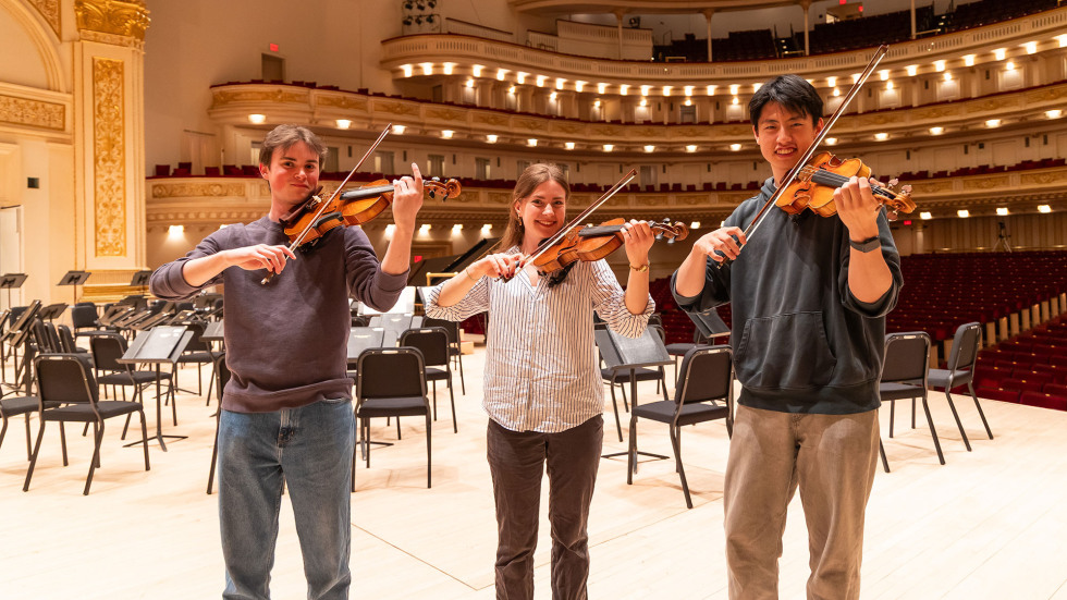 Three orchestra members stand on stage at Carnegie Hall holding violins.