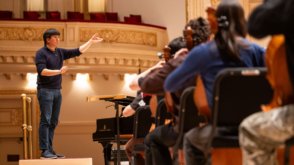 Mark standing on a platform conducting musicians who are seated in front of him. 