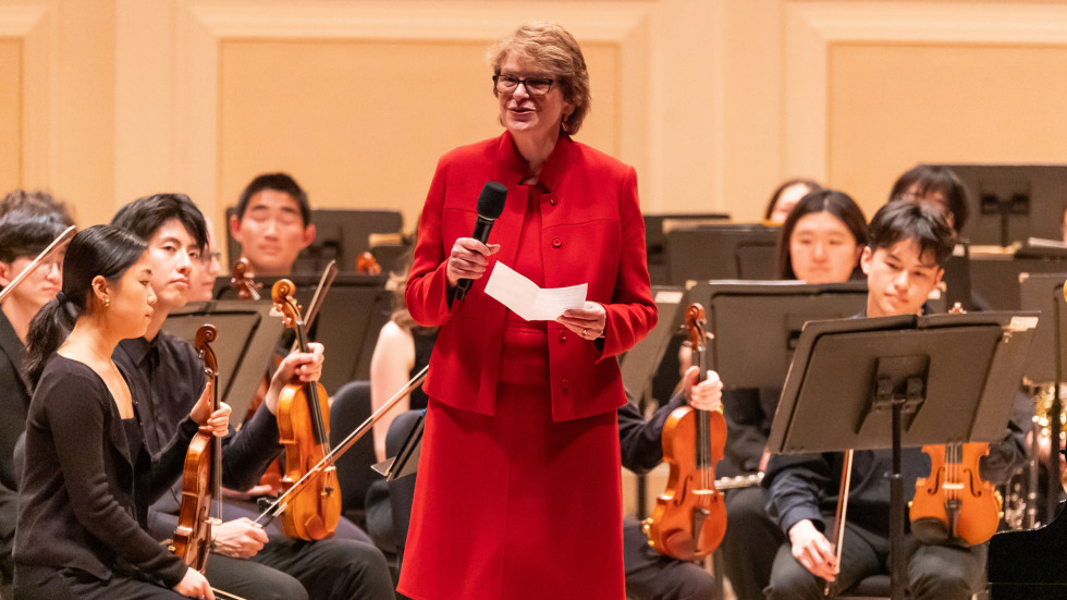 President Paxson speaking into a microphone on stage while the orchestra is seated behind her. 