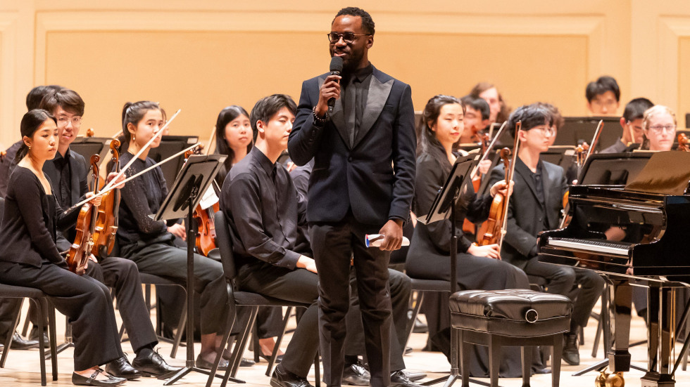 Joel stands on stage holding a microphone in front of the orchestra ahead of their performance. 