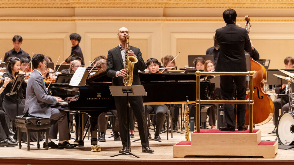 Brown Orchestra performing with pianist and saxophonist while conductor stands on a podium, leading the performance.