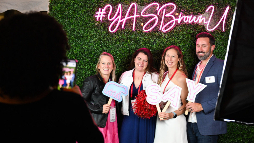 A group of young alums posing at the photo backdrop that says "YASBrownU"