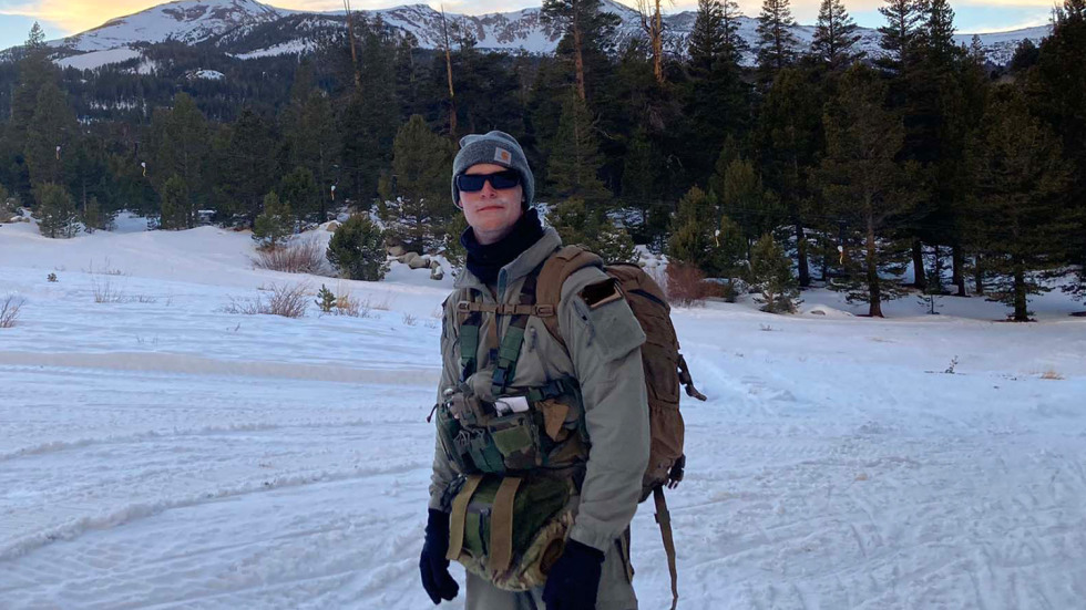 Tyler Hunter is wearing military gear and standing in on a snowy slope with mountains in the background.