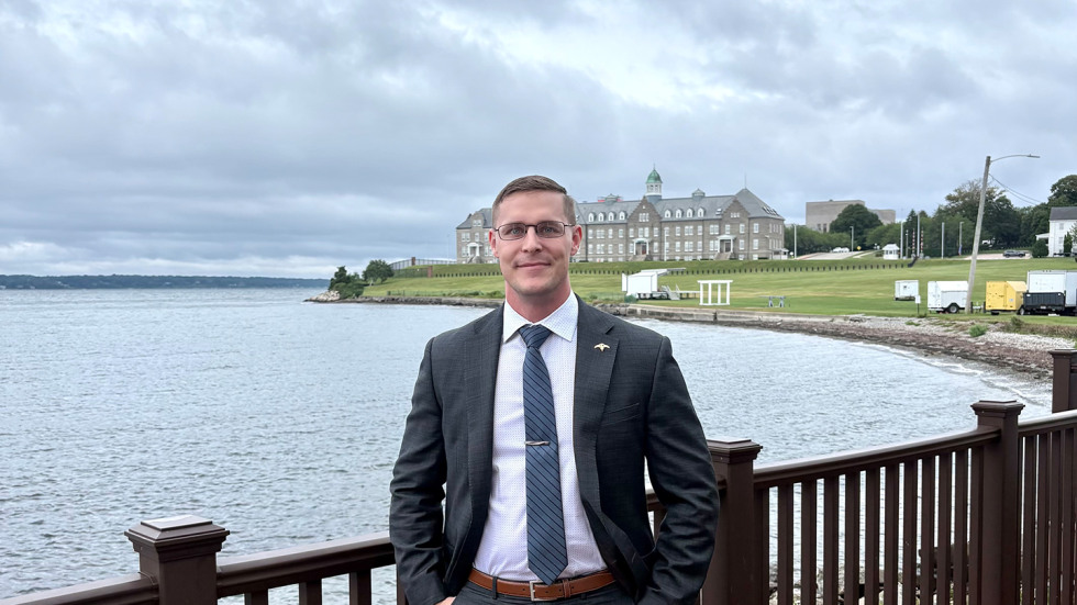 Tyler Hunter is wearing a suit and posing for the photo on a deck with water in the background.