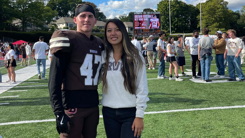 Wyatt Feltner is wearing a Brown football uniform, standing next to his wife. They're on the football field and other students are gathered in the background.